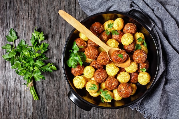 Albóndigas y bolas de espagueti horneadas en un plato negro para hornear - cocina italiana, servidas en un fondo de madera oscura con perejil fresco y cuchara de madera, vista superior, primer plano