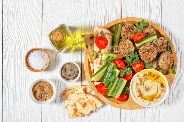 Albóndigas de cordero servidas con pan plano, pepino fresco, tomates y hummus en un plato de bambú sobre una mesa de madera blanca, plano, espacio libre