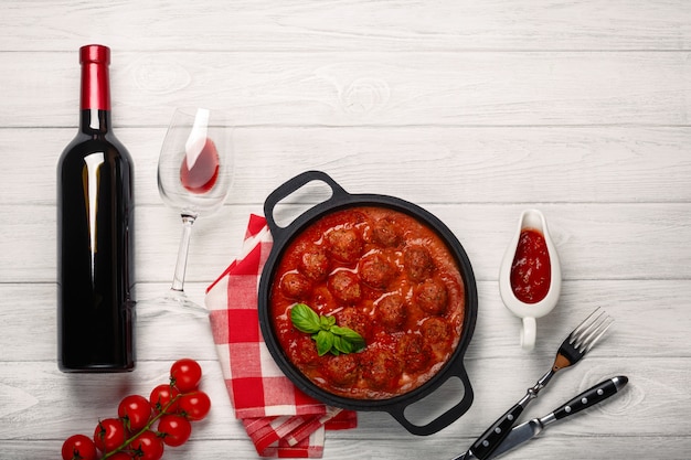 Albóndigas en salsa de tomate en una sartén con botella de vino, dos vasos, cuchillo y tenedor sobre una tabla de madera blanca