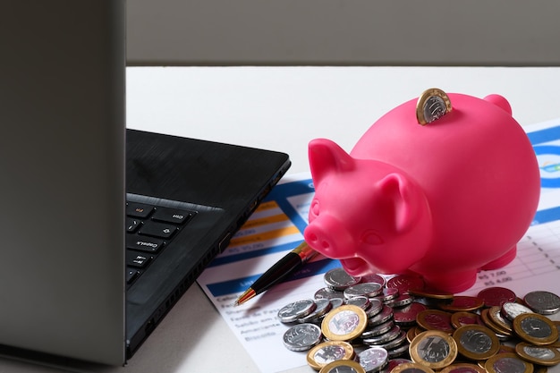 Una alcancía rosa se sienta encima de una computadora portátil junto a una pila de monedas bazilianas