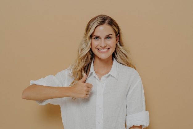 Alcanzando objetivos. Retrato de mujer joven positiva con amplia sonrisa mostrando el pulgar hacia arriba gesto con la mano mientras posa en el estudio contra la pared beige, mirando a la cámara con expresión feliz. Lenguaje corporal