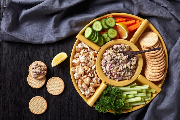 Alcaparras de atún encurtidos para untar servido con zanahorias y palitos de apio, rodajas de pepino fresco, galletas saladas y chicharrones en tazones sobre una mesa de madera oscura, cocina filipina, plano, espacio libre