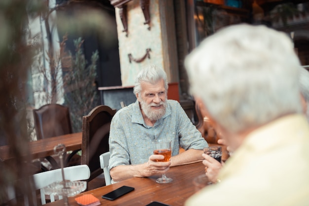 Alcohol con amigos. Hombre canoso con barba bebiendo alcohol con amigos mientras está sentado afuera