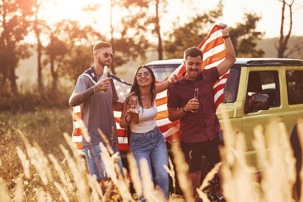 Con alcohol en las manos. Los amigos tienen un buen fin de semana al aire libre cerca de su coche verde con la bandera de Estados Unidos.