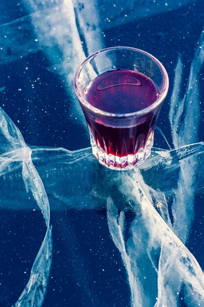 Alcohol en un vaso y hielo limpio con hermosas grietas profundas. Un vaso con tintura roja descansa sobre el hielo transparente del lago. Vertical.