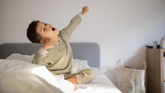 El alegre y activo niño caucásico despierta se sienta en la cama estirando el cuerpo y bosteza en el panorama interior del dormitorio