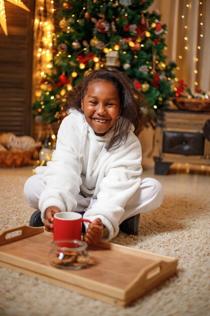 Alegre adolescente de piel oscura sosteniendo una taza roja con cacao en sus manos cerca del árbol de Navidad en casa concepción de Navidad