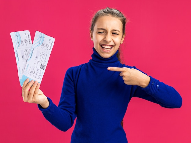 Alegre adolescente sosteniendo apuntando y mirando billetes de avión aislados en la pared rosa