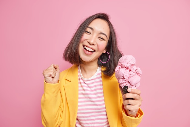 Alegre adorable adolescente con apariencia oriental, cabello oscuro, inclina la cabeza, aprieta el puño, come sabroso helado, sonríe ampliamente, se divierte disfruta del verano vestida con ropa elegante aislada en la pared rosa