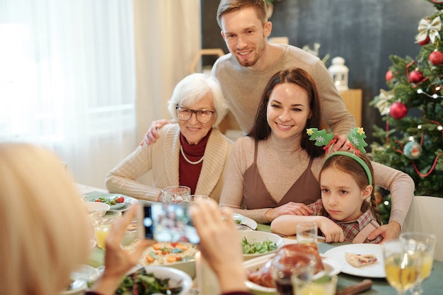 Alegre afectuosa familia sentada junto a la mesa festiva servida y mirando el teléfono inteligente en manos de una mujer madura