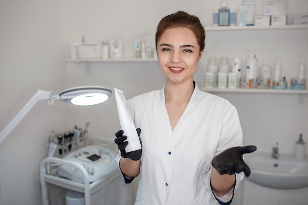 Alegre agradable esteticista de pie en la sala de cosmetología y sostener tubo crema. Ella mira y sonríe.