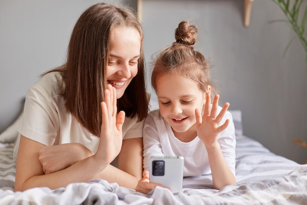 Alegre alegre mãe e filha usando telefone celular relaxando na cama em casa olhando para a tela do smartphone com videochamada acenando com a mão dizendo olá