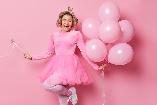 La alegre y alegre princesa viste vestidos festivos, baila despreocupada, sostiene una varita mágica y un montón de globos de helio inflados se divierten en una fiesta aislada sobre el fondo rosa del estudio. Toma monocromática.