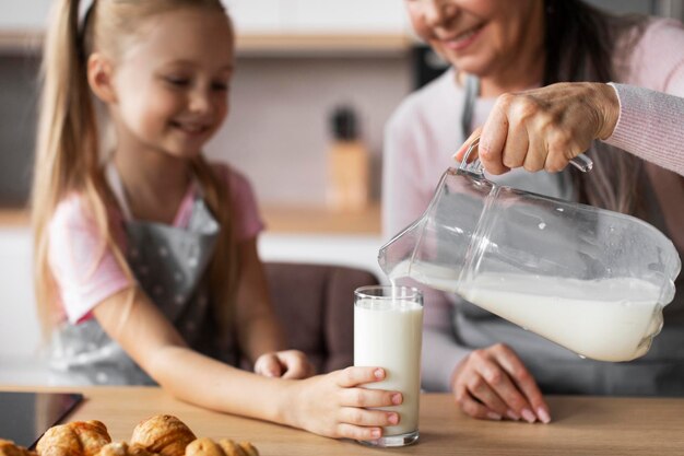 Alegre anciana caucásica vierte leche en un vaso para galletas frescas para que las niñas disfruten horneando