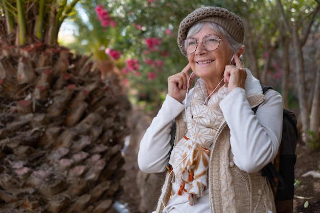 Foto alegre anciana con gorra y mochila en una excursión al aire libre sentada escuchando música con auriculares anciana atractiva descansando a la sombra de los bosques tropicales