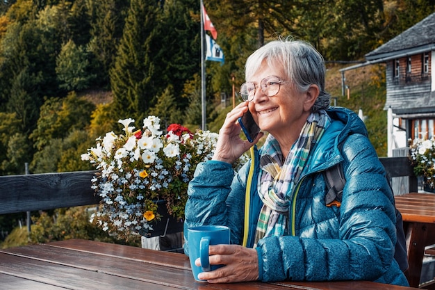 Alegre anciana sentada en la mesa del restaurante al aire libre en la montaña hablando por teléfono móvil mientras sostiene una taza de café Anciana viajera disfrutando de la libertad y la jubilación