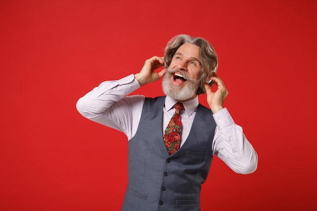 Alegre anciano canoso bigote barbudo hombre en camisa clásica chaleco corbata posando aislado sobre fondo rojo. Concepto de estilo de vida de las personas. Simulacros de espacio de copia. Escuche música con auriculares, mirando a un lado.