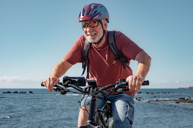 Alegre anciano caucásico con casco montando en la playa con bicicleta eléctrica, feliz jubilado