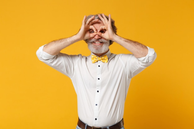 Alegre anciano de pelo gris bigote barbudo hombre con camisa blanca y pajarita posando aislado sobre fondo amarillo naranja. Concepto de estilo de vida de las personas. Simulacros de espacio de copia. Imitando anteojos o binoculares.