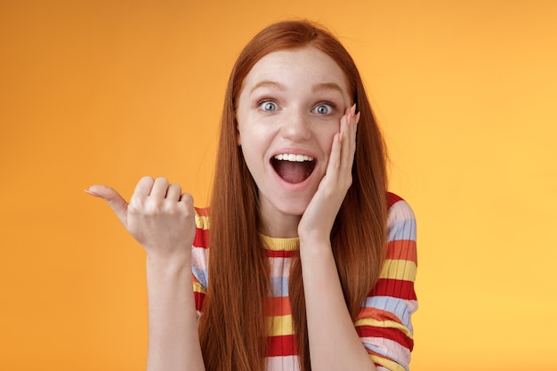 Alegre, animado, impressionado, falante, apontando para o polegar espantado, mandíbula esquerda, sorrindo amplamente, tocar a bochecha emocionada, evento interessante discutir desempenho maravilhoso incrível, fundo laranja.