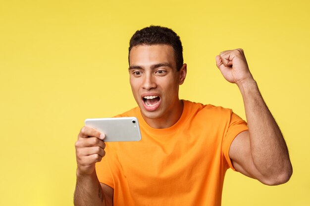 Alegre animado jovem bonito de camiseta laranja, levante a mão a bomba do punho como torcendo pelo time favorito, segurando o smartphone horizontalmente, assistindo futebol no dispositivo móvel, fundo amarelo