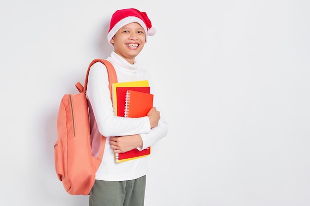 Alegre y apuesto joven estudiante asiático con un sombrero de Navidad usando una camiseta con mochila sosteniendo un libro aislado de fondo blanco