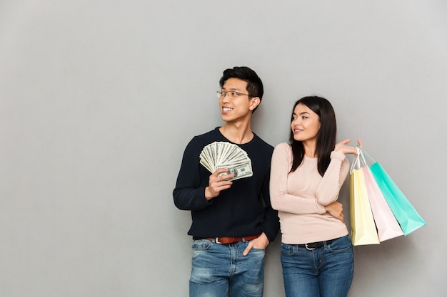 Alegre asiático jovem casal apaixonado, segurando o dinheiro e sacolas de compras. Olhando de lado.