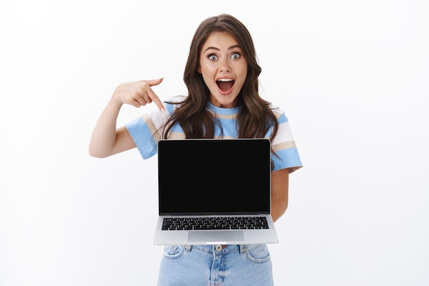 Foto alegre asombrada hermosa mujer caucásica sostenga la computadora portátil, deje caer la mandíbula fascinada señalando la pantalla de la computadora, mostrando la aplicación, tienda en línea en la pantalla del dispositivo, recomienda espacio de copia fresco, pared blanca