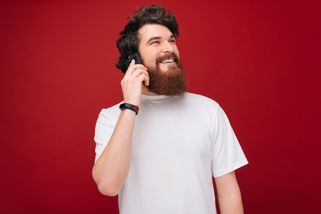 Alegre barbudo bonitão na camiseta branca falando no telefone sobre parede vermelha