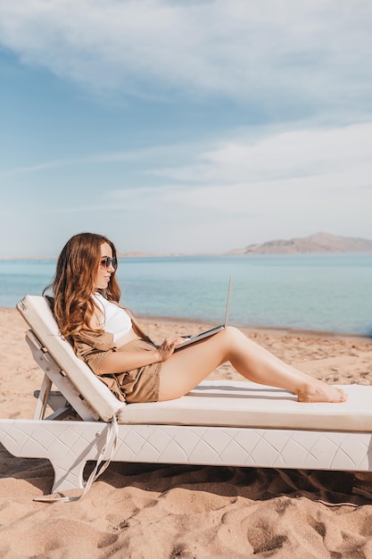 Una alegre bloguera trabaja de forma remota con una computadora portátil en la playa con vistas al mar Una mujer hace un video de un viaje Viaja por el mundo y trabaja con redes sociales en línea Profesión moderna remota