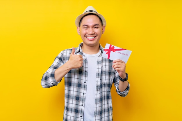 Alegre bonito etnia asiática jovem vestindo roupas casuais chapéu segurando vale-presente cupom vale aparecendo polegar como gesto isolado no fundo amarelo Pessoas conceito de estilo de vida