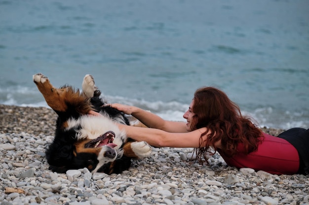 Alegre caminhada feliz e descanso com cachorro grande na manhã quente de verão Mulher caucasiana ruiva bonita coça a barriga de Bernese Mountain Dog e ri alegremente e sinceramente