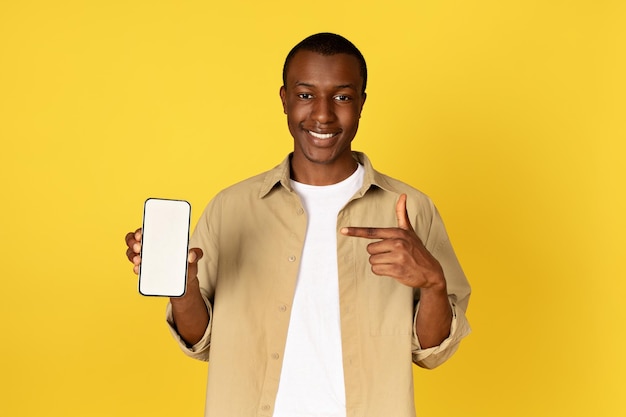 Alegre cara afro-americano milenar casual apontando o dedo para smartphone com tela em branco