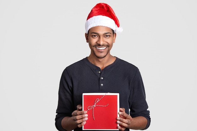 Alegre cara de pele escura tem sorriso, usa chapéu de Papai Noel, carrega caixa vermelha, vai fazer presente para a namorada no ano novo, interior de modelos, poses em branco.