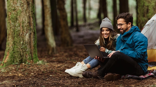 Alegre casal caucasiano milenar bebe café quente na floresta fria desfruta de aventura sentado na tenda digitando no laptop