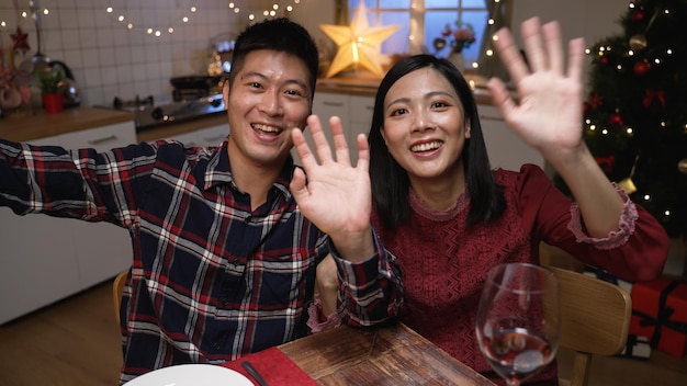 alegre casal japonês acenando oi e apontando para a câmera do celular enquanto faz videochamada para dar cumprimentos de ano novo ao amigo no jantar em um interior de casa festivo
