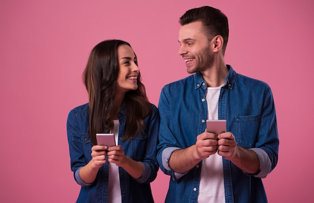 Alegre casal jovem isolado sobre fundo rosa, segurando telefones inteligentes em roupas casuais