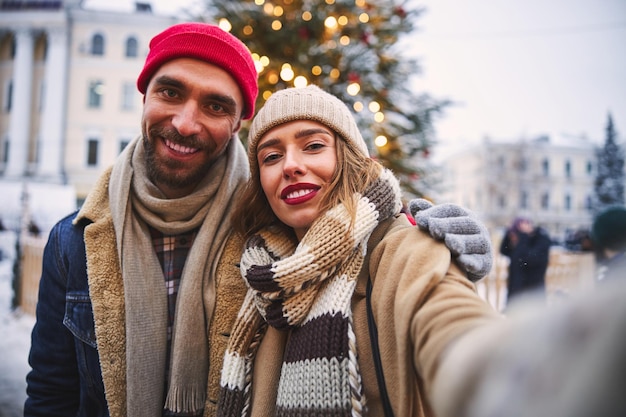 Alegre casal jovem tomando selfie na árvore de Natal ao ar livre