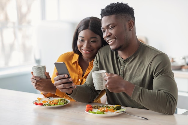 Alegre casal negro assistindo vídeos engraçados no smartphone durante o café da manhã