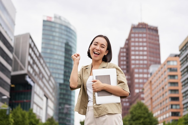 Alegre chica asiática en el centro de la ciudad mostrando un gesto de sí hurra de pie con una computadora portátil y celebrando