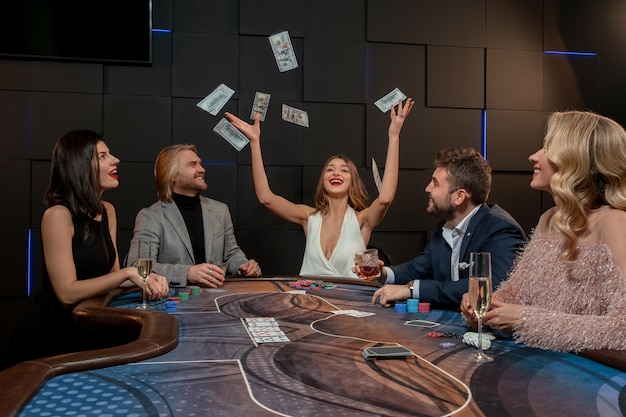 Alegre chica de cabello castaño rompiendo el banco en un juego de póquer, lanzando billetes al aire mientras se sienta en la mesa de juego con los oponentes. Concepto de juego de buena suerte