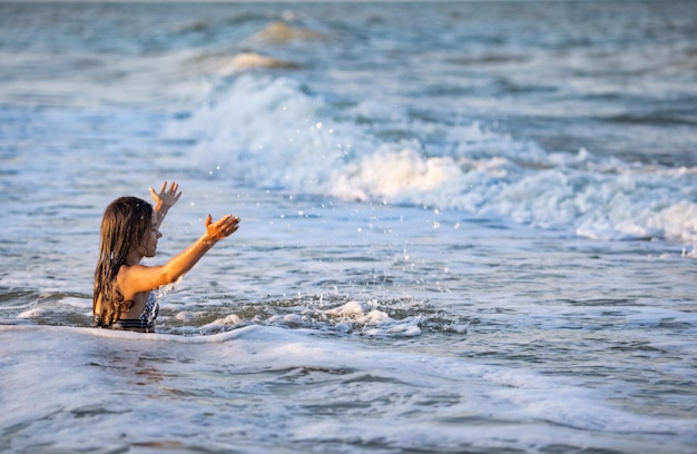 Alegre chica mojada de pelo oscuro con el pelo mojado en un traje de baño de bikini de leopardo brillante salpica agua a su alrededor y disfruta del cálido sol mientras se sienta en el mar azul de verano