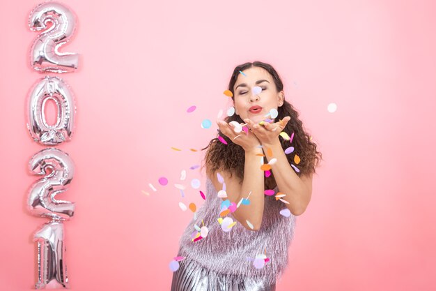 Alegre chica morena elegante con el pelo rizado en ropa festiva lanza confeti en una pared rosa con globos plateados a la izquierda para el concepto de año nuevo