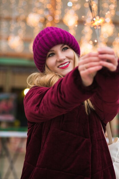 Alegre chica rubia vestida con ropa elegante, sosteniendo bengalas brillantes en la feria de Navidad en Kiev