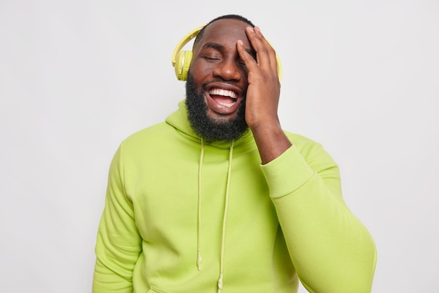 un alegre chico barbudo de piel oscura mantiene la mano en la cara ríe felizmente escucha música en auriculares disfruta del tiempo libre en un pasatiempo vestido con una sudadera con capucha verde posa en blanco disfruta de un buen sonido