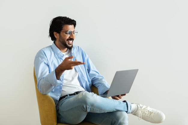 Alegre chico milenario del medio oriente con gafas señala con el dedo a la computadora portátil sentada en el sillón tiene un video