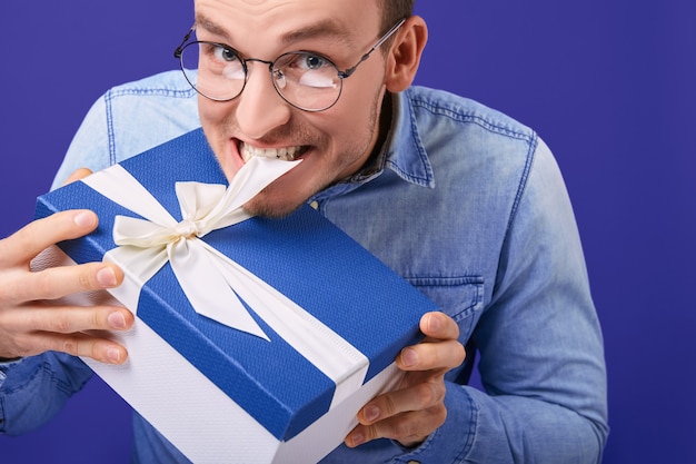 Alegre chico sonriente con gafas redondas tratando de desatar el regalo con los dientes