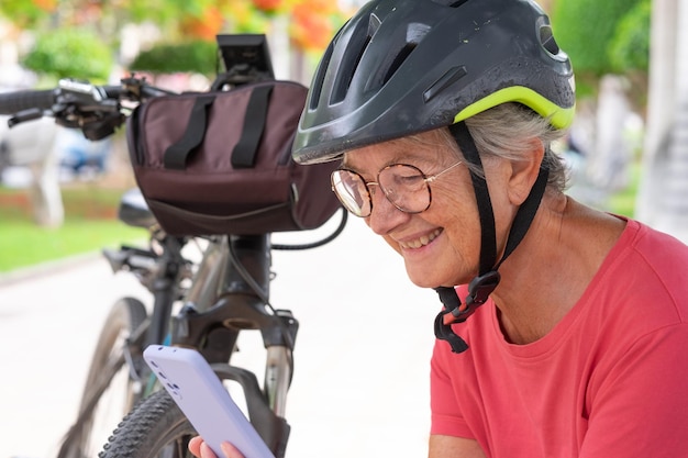 Alegre ciclista anciana con casco y anteojos sentada en un banco en un parque público cerca de su bicicleta usando un teléfono móvil leyendo un mensaje