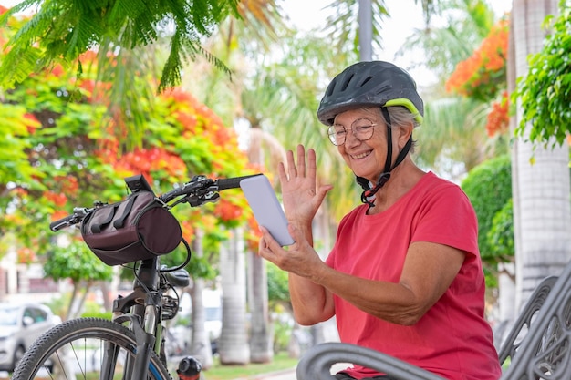 Alegre ciclista senior mujer sentada en un banco en un parque público cerca de su bicicleta en video chat por teléfono móvil