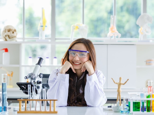 Alegre científica étnica en gafas protectoras sentada a la mesa con frascos de química y microscopio en el laboratorio mientras se apoya en la mano y mira a la cámara